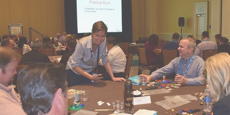 Annemarie Boss stands next to a table of people as she facilitates the group in a LEGO® Serious Play exercise.